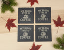 Four dark gray coasters with 'My blood type is coffee' text on a wooden surface with autumn leaves.