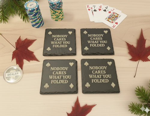 Four black slate coasters with humorous text on a wooden surface.