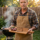 Man holding a wooden cutting board with 'Dad's BBQ' engraving, standing in a backyard with a grill.