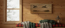 A rustic wooden wall art piece depicting two ducks in flight, displayed on a wood-paneled wall above a red plaid bench with knitted blankets and a lantern.