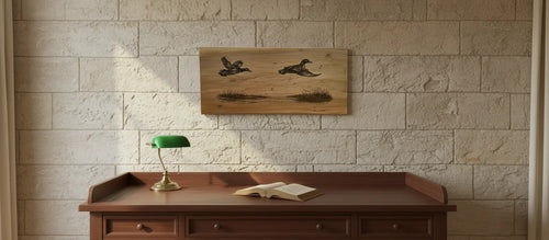 A rectangular wooden wall art piece featuring two flying ducks and a duck on a lake, displayed on a light stone brick wall above a dark wood desk with a green lamp and an open book.