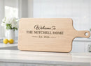 Wooden cutting board with engraved text 'Welcome To THE MITCHELL HOME Est. 2026' on a kitchen counter.