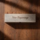 A rectangular light gray wooden box engraved with the words 'New Beginnings' in dark script, placed on a rustic brown wooden surface with soft shadows.