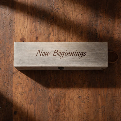 A rectangular light gray wooden box engraved with the words 'New Beginnings' in dark script, placed on a rustic brown wooden surface with soft shadows.