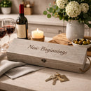 A rustic wooden gift box engraved with 'New Beginnings' sits on a kitchen counter next to a wine bottle, two wine glasses, a candle, keys, and a vase of flowers.