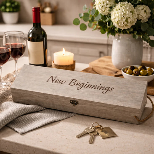 A rustic wooden gift box engraved with 'New Beginnings' sits on a kitchen counter next to a wine bottle, two wine glasses, a candle, keys, and a vase of flowers.