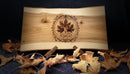 A rectangular live-edge wood board with a natural maple leaf design burned into the center, displayed on a stand with scattered autumn leaves in the foreground against a dark background.