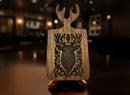 A rectangular wooden cutting board with a deer head and antlers design engraved on the front, displayed on a small stand on a dark wooden table with a blurred bar or kitchen background.