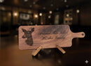 A personalized wooden serving board or cutting board on a small stand, featuring a dark engraved deer head silhouette and the phrase 'Around This Table We Are Family' in script, set against a blurred warm-toned indoor background.