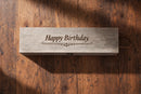 A rectangular wooden gift box engraved with 'Happy Birthday' and a small decorative flourish, placed on a rustic dark wood surface.